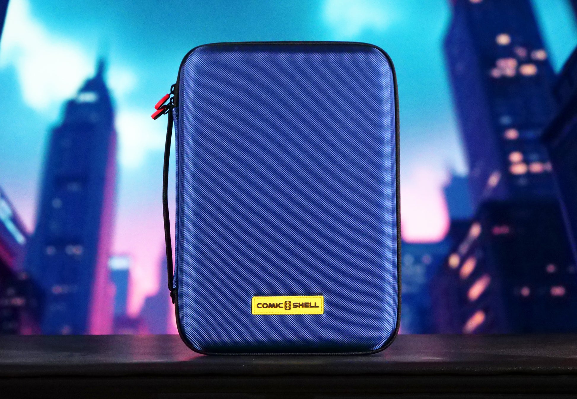 Blue Comic Shell graded comic travel case with a yellow logo on a blurred cityscape background